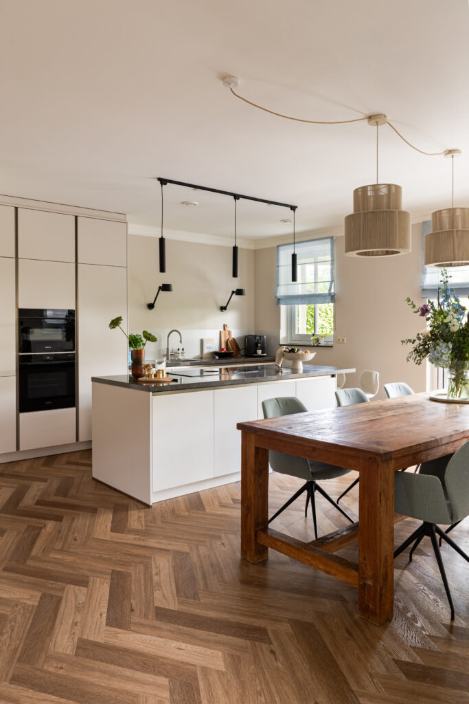 Open keuken met angora grijs keukeneiland en donker keramisch blad, robuuste houten eettafel en saliegroen stoelen — modern landelijk interieur met visgraatpvc,  hanglampen en zwarte inbouwapparatuur.