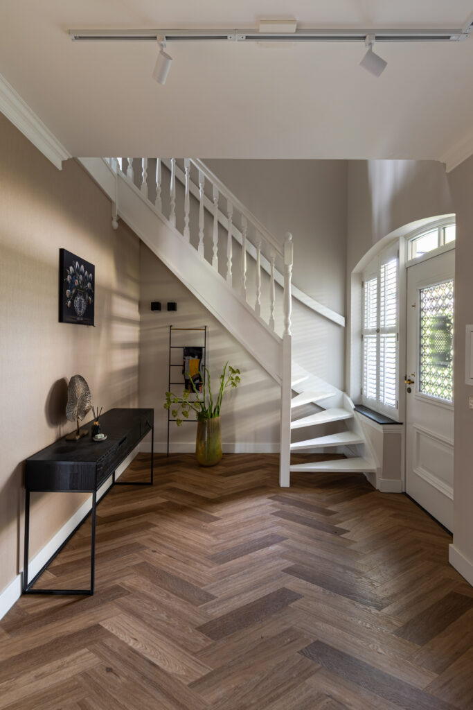 Klassieke entrée met visgraatparket, witte houten trap en originele glas-in-looddeur — stijlvolle hal met zwart sidetable, gouden decoratie en natuurlijk licht via authentiek boogvenster.
