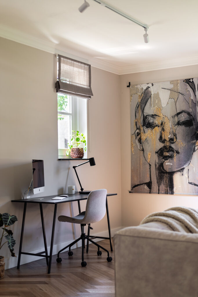 Stijlvolle thuiswerkplek met zwart bureau, grijze bureaustoel en iMac, gecombineerd met een groot abstract wandkleed met vrouwenportret in grijs en goud — lichte werkkamer met visgraatparket en linnen vouwgordijn.