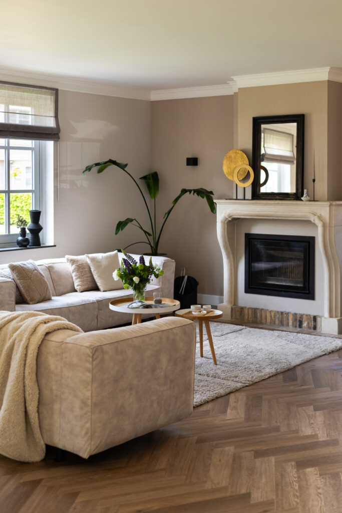 Moderne klassieke landelijke woonkamer met visgraatpvc, beige hoekbank, witte schouw en decoratief olifanten wandkleed — warm interieur met natuurlijke materialen en gouden accenten.