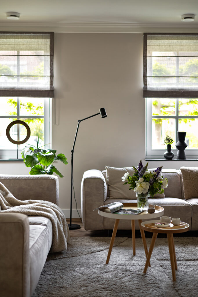 Lichte woonkamer met beige hoekbank, ronde houten salontafels en vers bloemstuk — sfeervol interieur met zacht vloerkleed, zwarte staande lamp en natuurlijk licht via linnen vouwgordijnen.