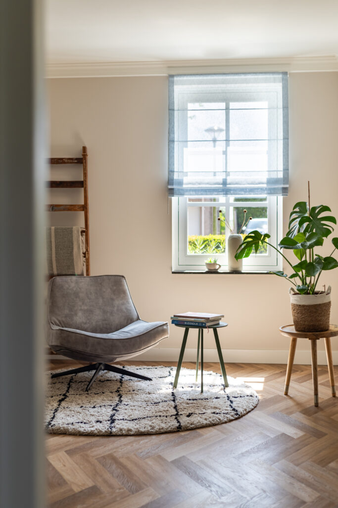 Sfeervolle leeshoek in de woonkeuken met grijze draaifauteuil op rond hoogpolig vloerkleed en visgraatparket — rustige zitplek met monstera, houten decorladder en natuurlijk licht via blauw vouwgordijn.