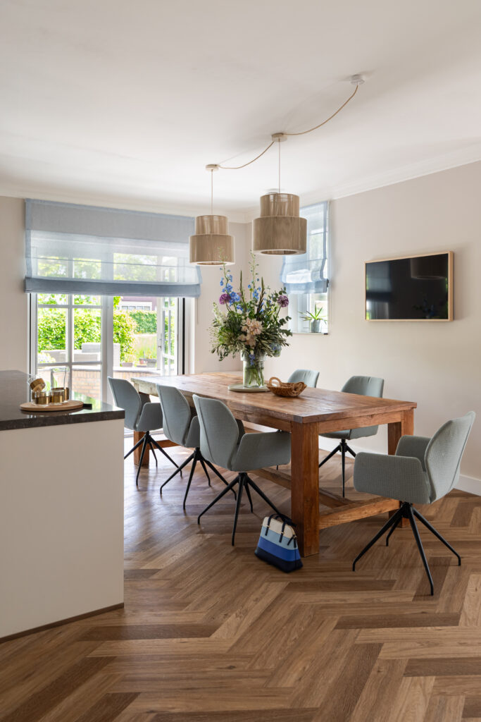 Lichte eetkamer met robuuste houten eettafel, licht blauwe stoelen en hanglampen — modern landelijk interieur met visgraatpvc, vers bloemstuk en tuindeuren met doorkijk naar groene tuin.