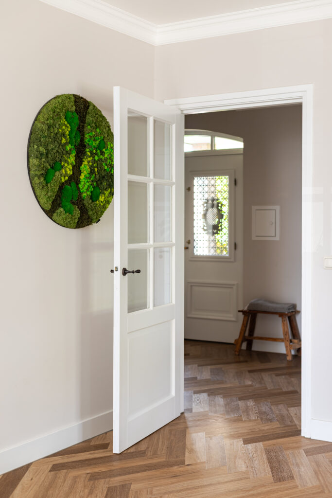 Lichte hal met visgraatparket, witte glasdeur en rond mosschild als wanddecoratie — klassiek interieur met doorkijk naar entrée met originele glas-in-looddeur en houten krukje.