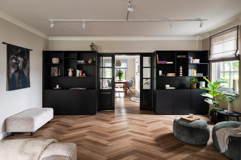Ruime woonkamer met visgraatparket en twee grote zwarte boekenwanden met stalen deuren — modern klassiek interieur met olifanten wandkleed, groene planten en doorkijk naar lichte eetkamer.