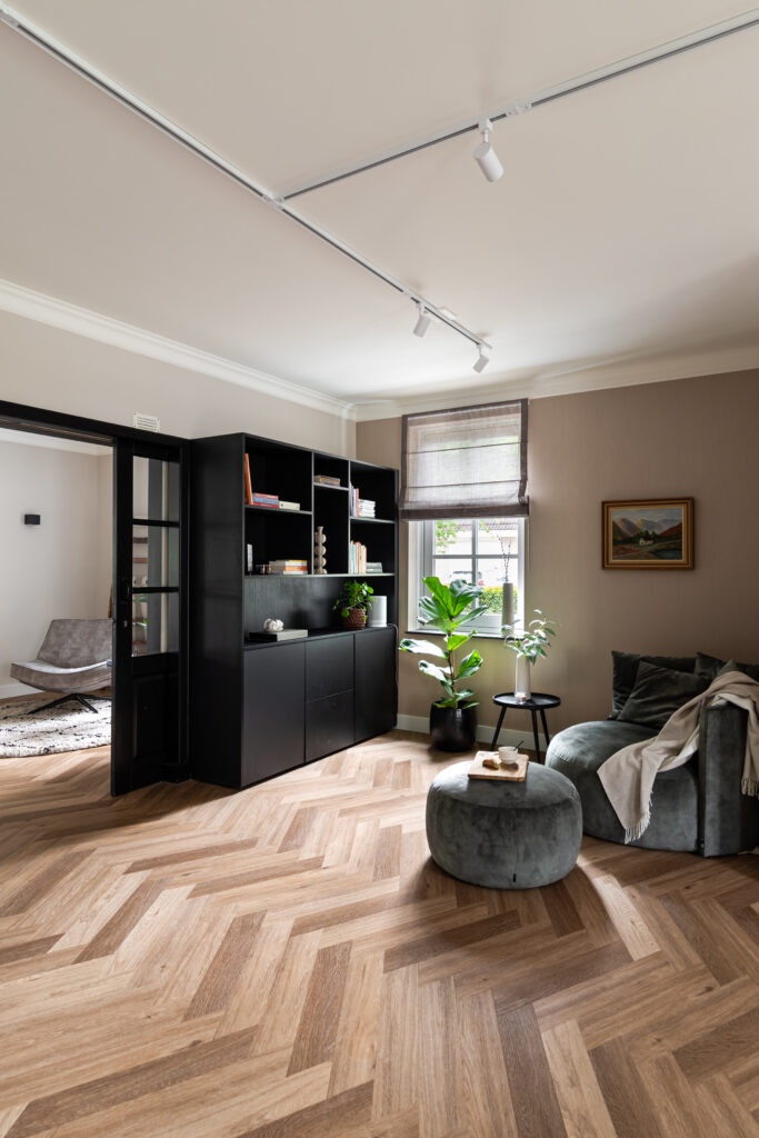 Moderne woonkamer met visgraatpvc, zwarte boekenkast en grijsgroene fauteuil met velvet poef — sfeervol interieur met taupe muren, groene planten en natuurlijk licht via linnen vouwgordijn. Leeshoek